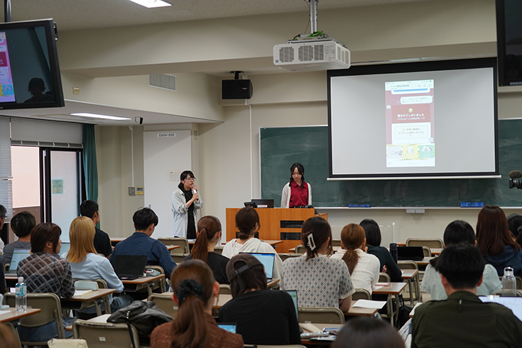 学生のスマートフォンで「ニセ警察詐欺仮想被害体験ツール」の操作説明する甲斐さん(左側)